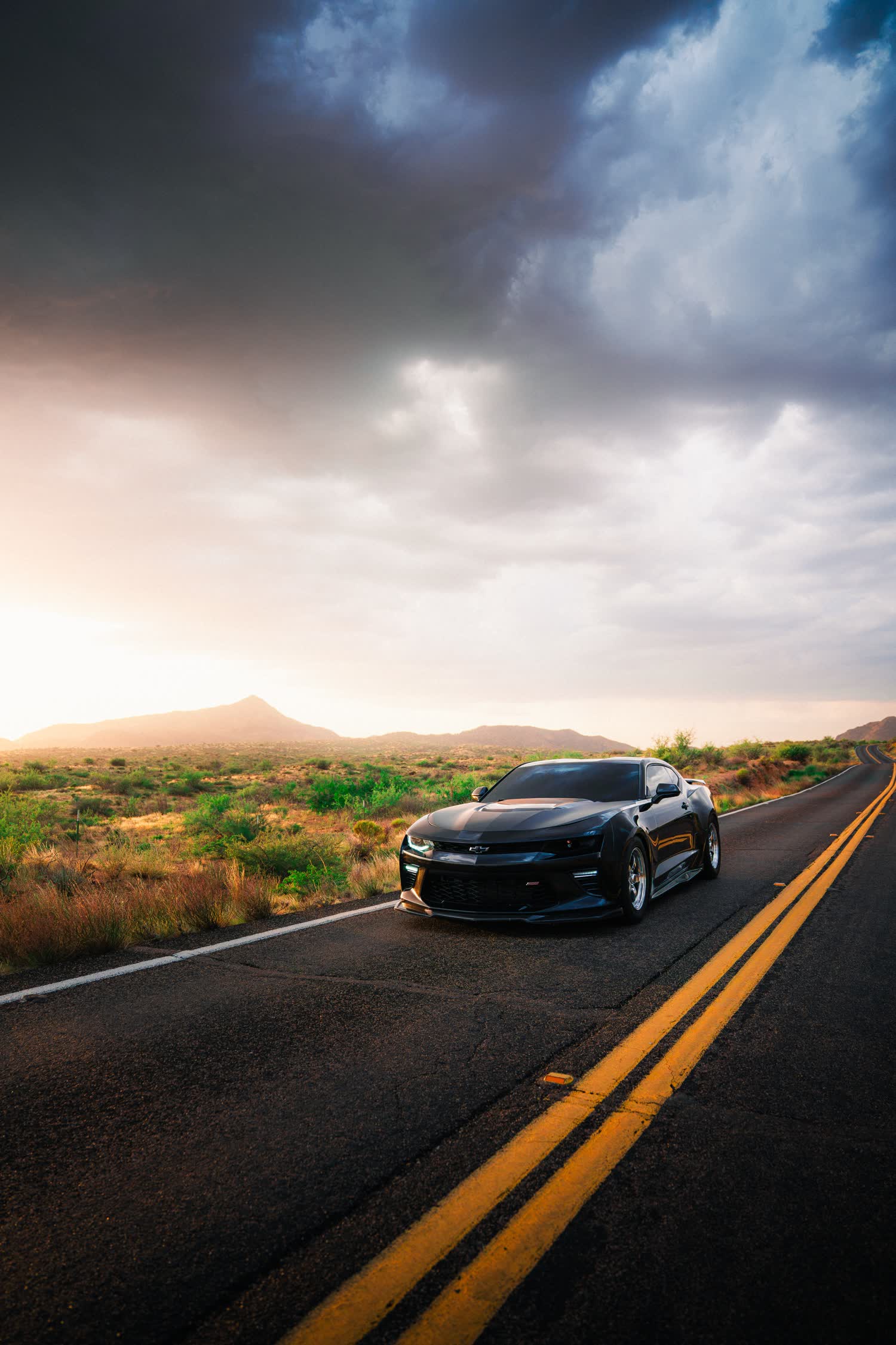 A widebody Camaro on a desert road at sunset, storm clouds above the mountains