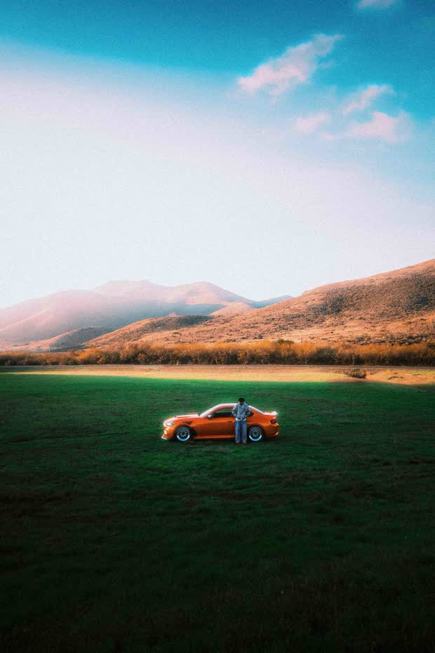 An orange coupe parked alone in a wide green field beneath golden hills