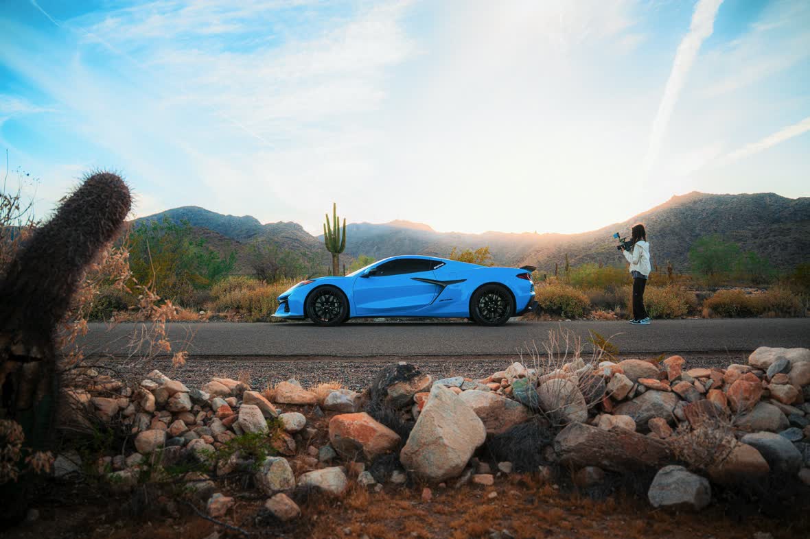 A blue Corvette on a desert road past saguaro cactus and mountains