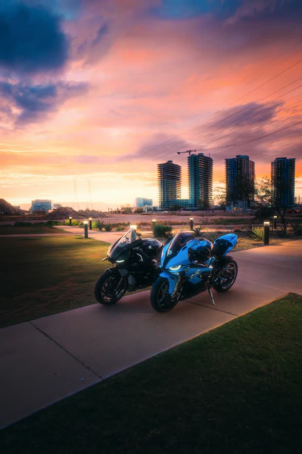 Two sportbikes parked on a path with a city skyline and coral sunset behind